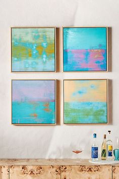 four abstract paintings hang on the wall above a dresser with bottles and glasses next to it