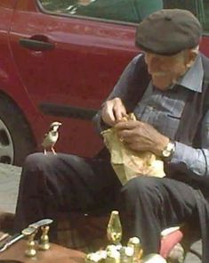 an old man sitting on the ground next to a red car