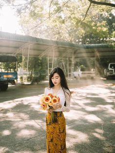 a woman holding flowers standing in front of a bus stop with the sun shining on her face