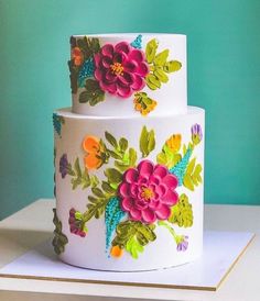 a three tiered cake decorated with flowers and leaves on a white table against a teal green background