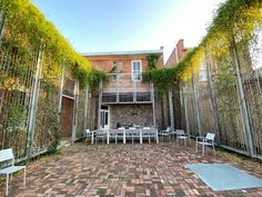 an outdoor dining area with white chairs and table surrounded by greenery on either side of the building