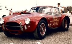 an old red sports car parked in a parking lot next to other cars and people