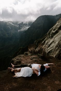 two people are laying on the ground in front of some mountains with clouds above them