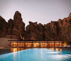 an outdoor swimming pool surrounded by mountains and rocks at dusk with lounge chairs around it