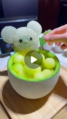 a person is holding a small teddy bear in a bowl filled with green apples and grapes