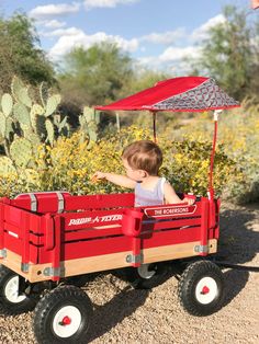 a little boy riding in a red wagon with an umbrella on it's roof