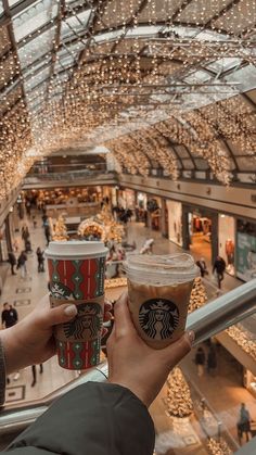 someone holding up two cups of starbucks coffee in a mall with lights hanging from the ceiling