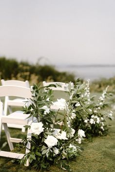 white chairs lined up on the grass with flowers and greenery in front of them