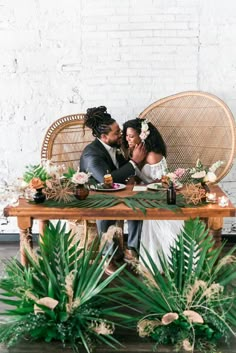 a bride and groom sitting at a table with greenery in front of their faces