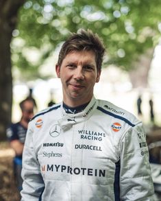 a man standing in front of a tree wearing a white racing suit and smiling at the camera