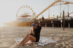 a woman sitting on top of a towel in front of a roller coaster at the beach