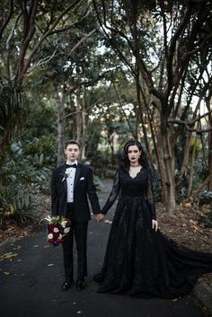 a man and woman in formal wear holding hands while standing on a path surrounded by trees