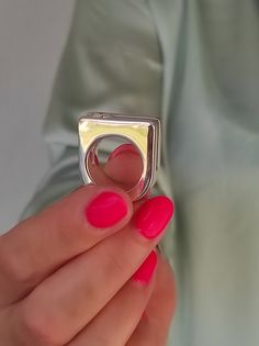 a woman's hand holding a ring that is shaped like a square, with pink nail polish on it