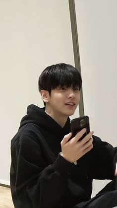 a young man sitting on the floor looking at his cell phone while wearing a black hoodie