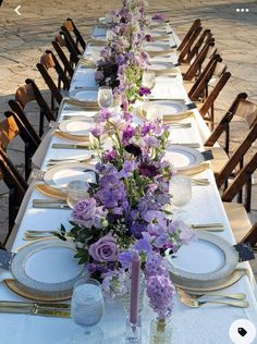 a long table is set with purple flowers and place settings for the guests to sit at