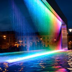 a rainbow fountain in the middle of a city at night with lights reflecting off it's water