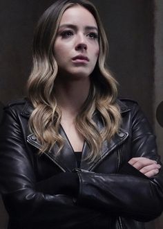 a woman with her arms crossed standing in front of a dark background, wearing a black leather jacket