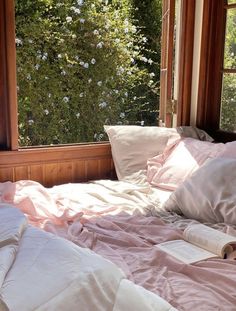 an unmade bed in front of a window with pink sheets and pillows on it