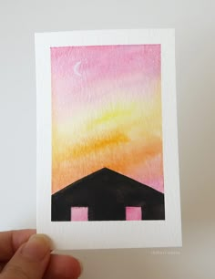 a hand holding up a watercolor painting of a sunset over a house with pink and yellow colors