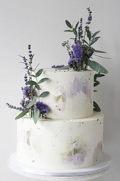 a three layer white cake with purple flowers and greenery on top, sitting on a plate