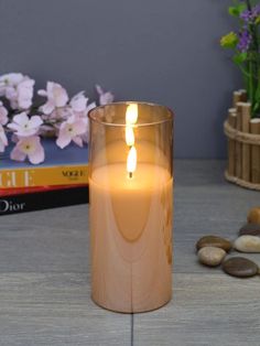 A lit candle in a glass holder sits on a wooden surface, surrounded by books, pink flowers, and smooth stones. The setting is calm and cozy.