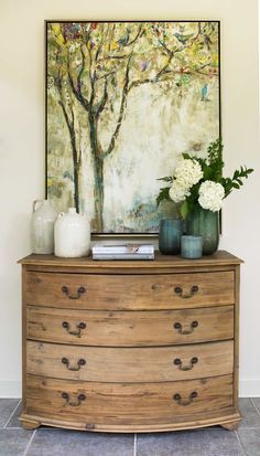 a dresser with flowers and vases on it in front of a large tree painting