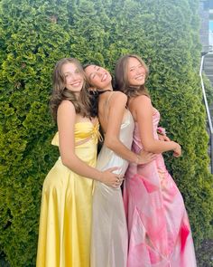 three young women in dresses standing next to each other near a green wall and bushes