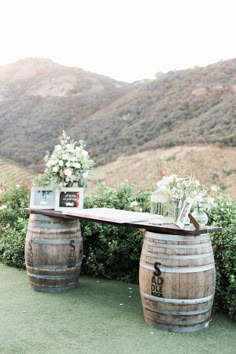 a wooden table topped with two wine barrels filled with flowers and greenery next to bushes