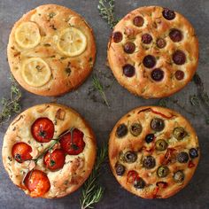 four pizzas with different toppings on top of a slate counter next to herbs and lemon slices