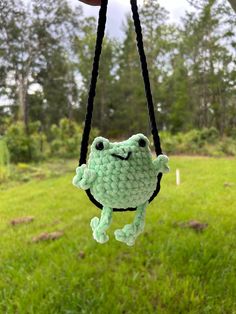 a green crocheted frog hanging from a black cord in the grass with trees in the background