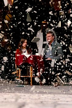 a man and woman sitting on a chair in front of snow falling from the sky