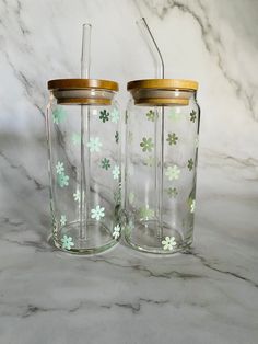 two clear glass jars with gold lids and green flowers on the sides, one has a straw in it