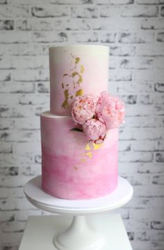 a three tiered cake with pink flowers on top and gold leaf decoration, sitting on a white pedestal