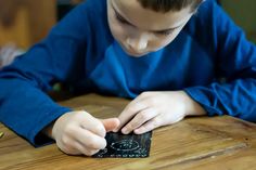 a young boy sitting at a wooden table playing with a piece of black paper on top of it