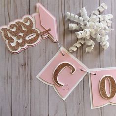 pink and gold birthday banner with the letter c hanging from it's side on a wooden table