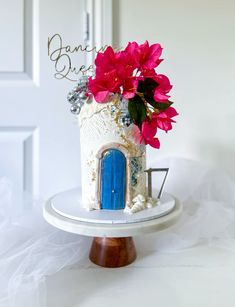 a white cake with blue door and pink flowers on top is sitting on a stand