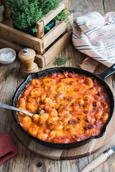 a skillet filled with pasta and sauce on top of a wooden table next to some herbs