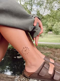 a woman's foot with a small compass tattoo on her left side leg, sitting next to a body of water