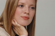 a woman with long blonde hair wearing a white shirt and looking at the camera while she has her hand on her chin