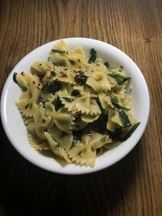 a white bowl filled with pasta and spinach on top of a wooden table next to a fork