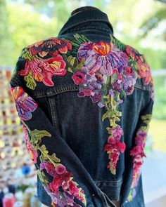 a woman's jean jacket with colorful flowers on the back and side, sitting on a table in front of a window