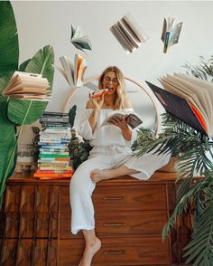 a woman sitting on top of a dresser holding a book and eating something in front of her face