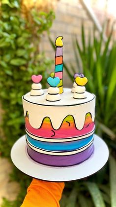 a hand holding up a cake with candles on it in front of some plants and bushes