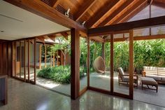 the inside of a house with glass walls and wood ceilinging, surrounded by greenery