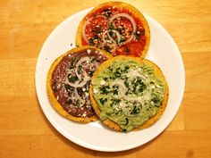 three small pizzas on a white plate topped with cheese and toppings sitting on a wooden table