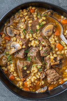 a bowl filled with meat and barley soup on top of a gray table next to a spoon