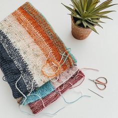 a pile of knitted blankets next to a potted plant and pair of scissors