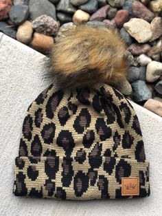 a leopard print beanie with a faux fur pom on the top, sitting next to some rocks