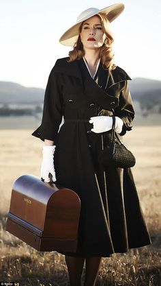 a woman in a black dress and hat holding a brown suitcase while standing in a field