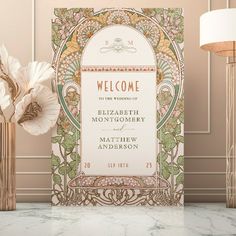 an ornate welcome card on a marble table next to a vase with flowers in it